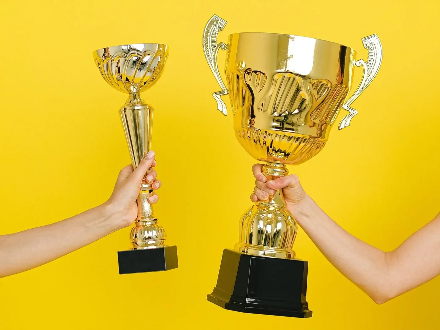 Two hands holding up gold trophies against a yellow background