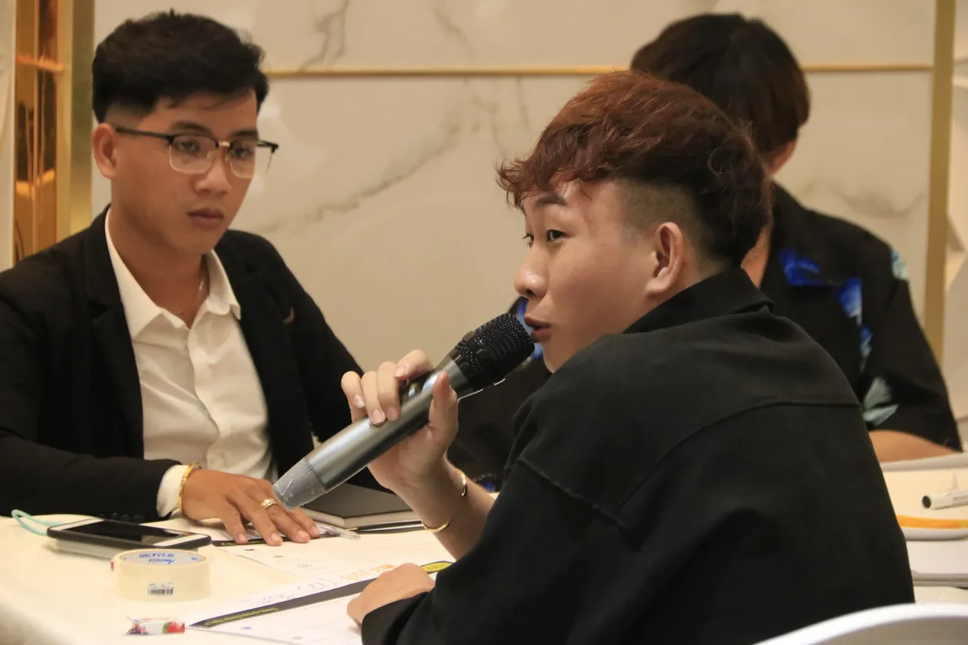 Two young men discussing ideas at a workshop table, one holding a microphone
